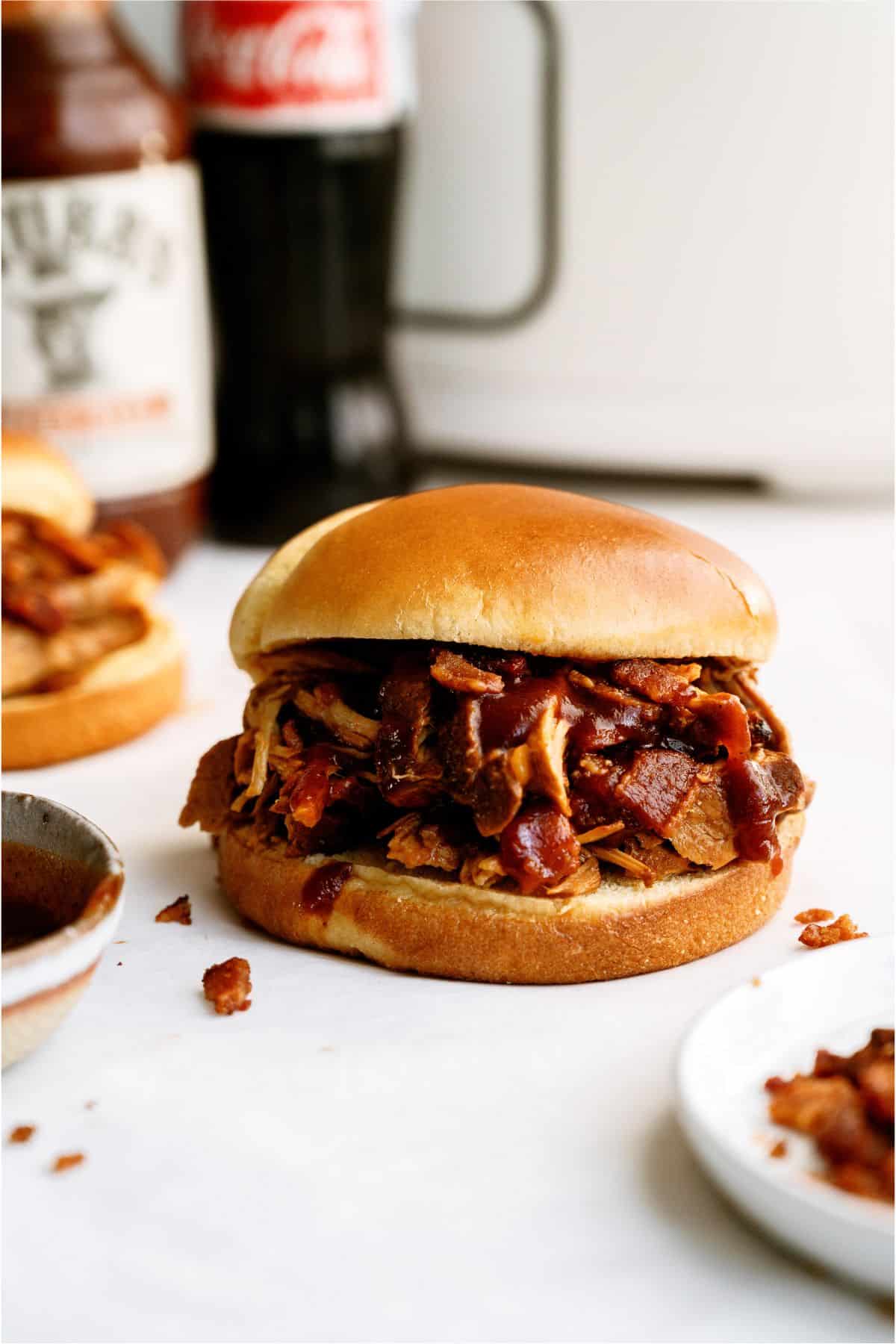 A pulled pork sandwich with barbecue sauce on a bun sits on a white surface, with a bottle of Coca-Cola and another sandwich in the background.