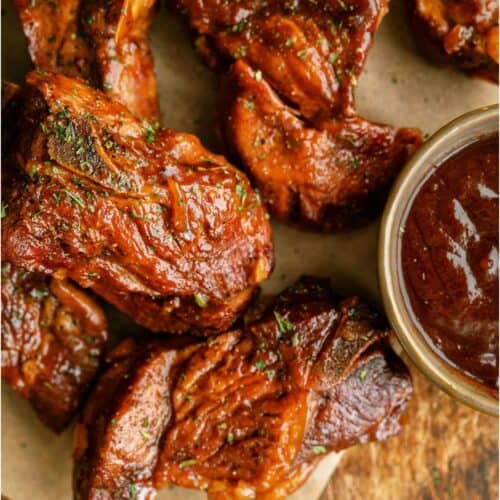 Several pieces of cooked, sauced pork country-style ribs are arranged on brown parchment paper beside a small bowl of barbecue sauce.