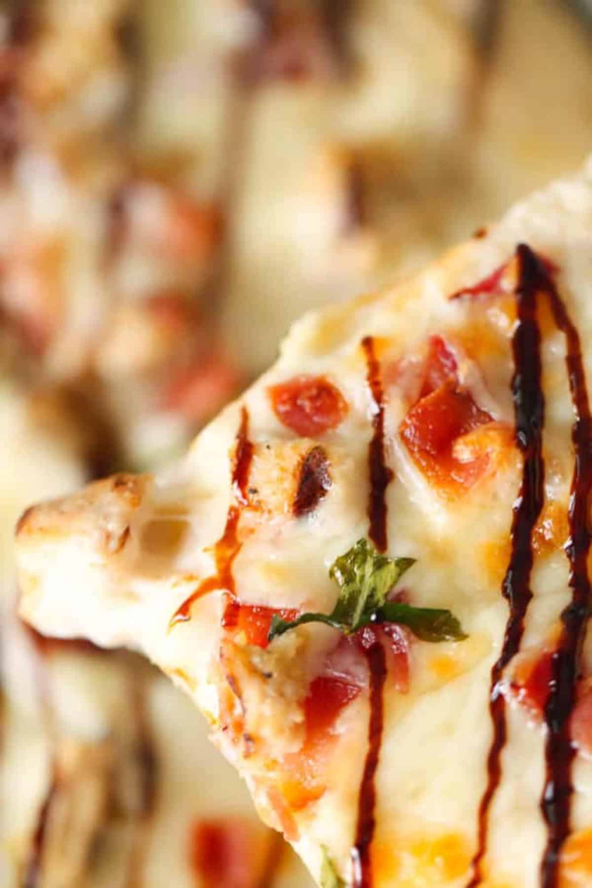 Close-up of a slice of pizza topped with melted cheese, diced tomatoes, grilled chicken, herbs, and a drizzle of balsamic glaze.