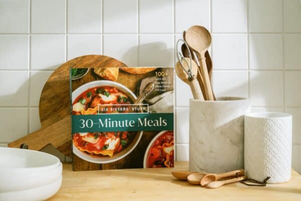 Feature image A cookbook titled "30-Minute Meals" stands on a kitchen counter beside wooden utensils in a marble holder and a white textured container.