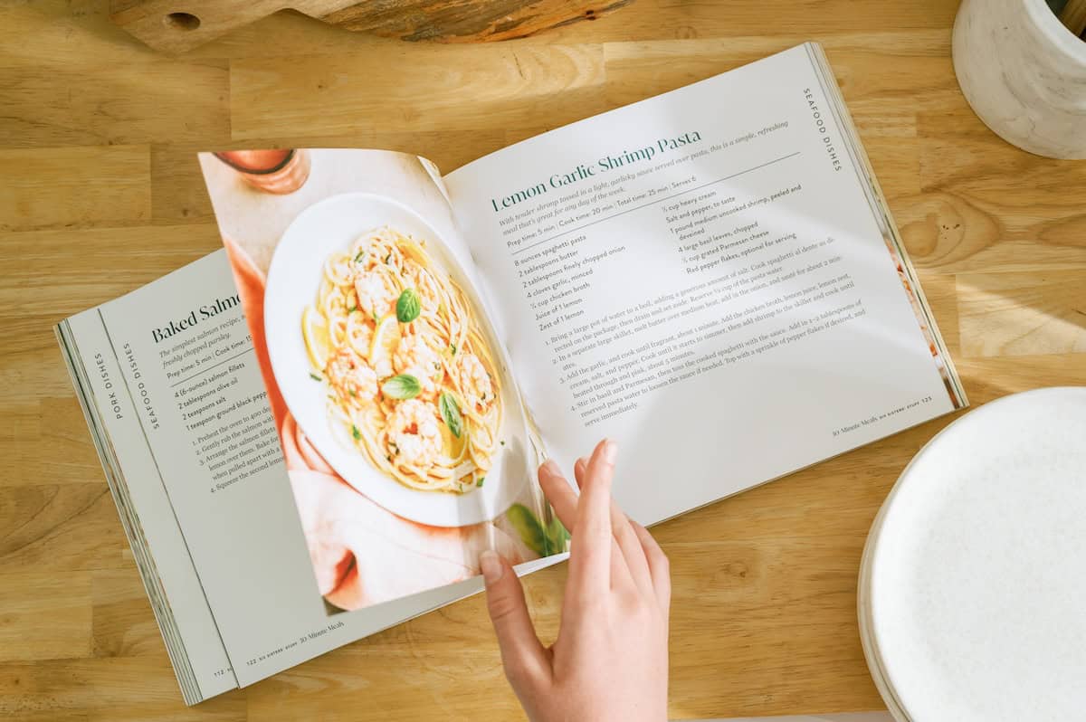 A hand turns the page of a cookbook opened to a recipe for Lemon Garlic Shrimp Pasta, with a photo of the dish on one page and text instructions on the other, on a wooden table.