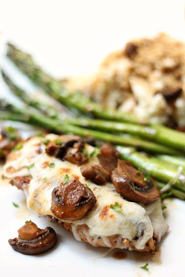 A plate of food featuring grilled chicken breast topped with melted cheese and sautéed mushrooms, accompanied by asparagus spears and a side of mashed potatoes.