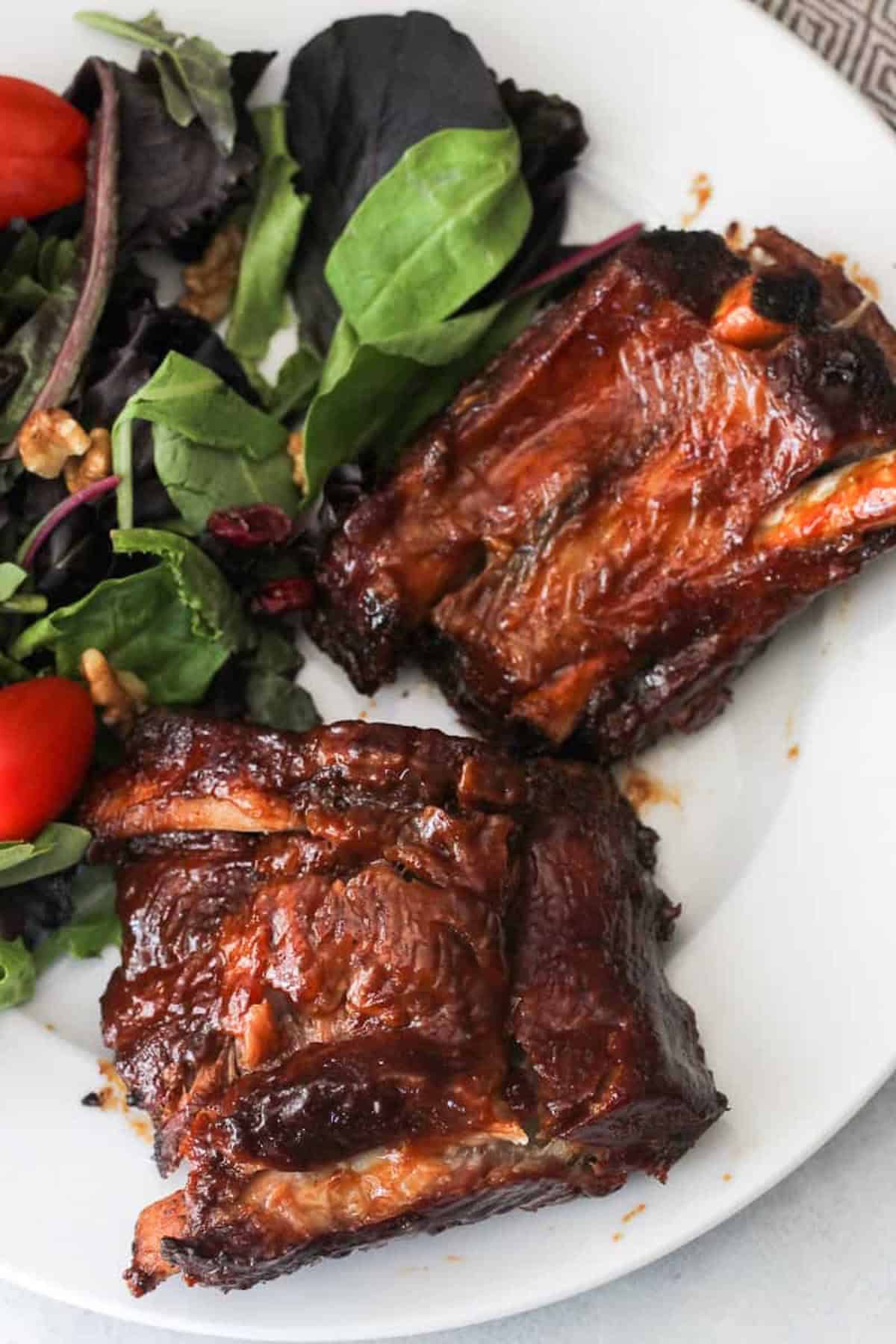 Two pieces of glazed barbecue ribs served on a white plate with a mixed green salad, grape tomatoes, and walnuts.