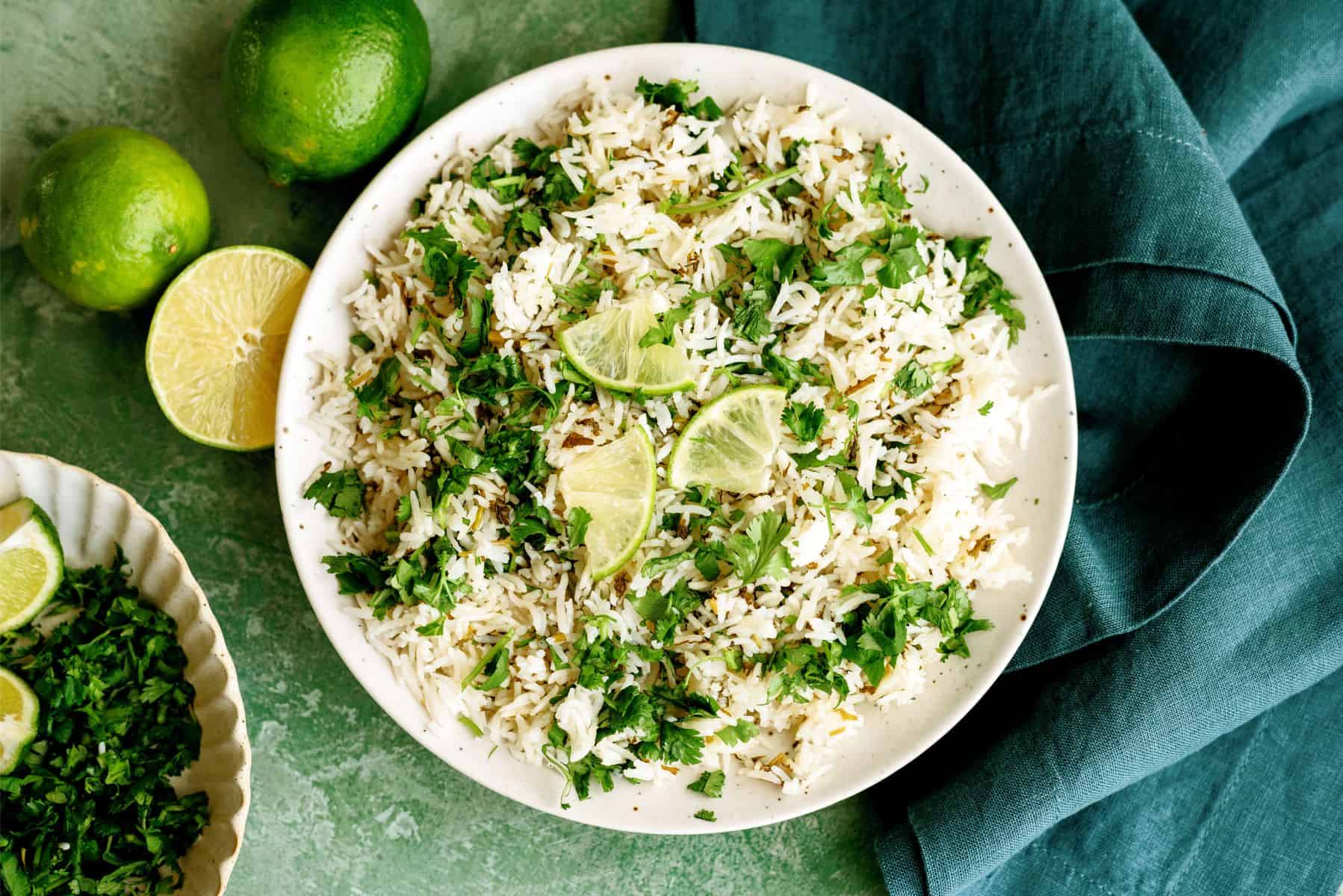 A bowl of white rice garnished with chopped cilantro and lime wedges, placed on a green surface with whole and sliced limes nearby.