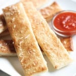 A square white plate holds several breadsticks, lightly dusted with cheese, served alongside a small dish of marinara sauce.