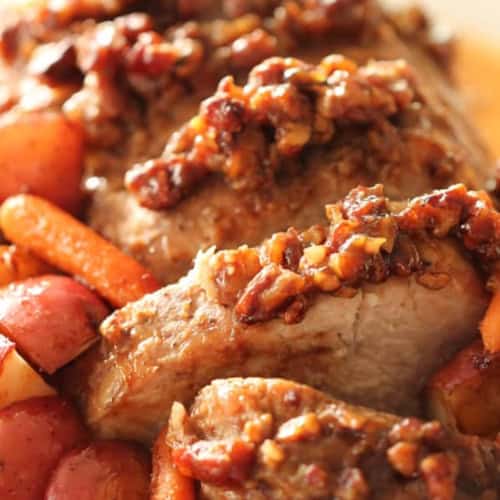 Sliced pork roast topped with a chopped nut glaze, served with cooked baby carrots and red potatoes.