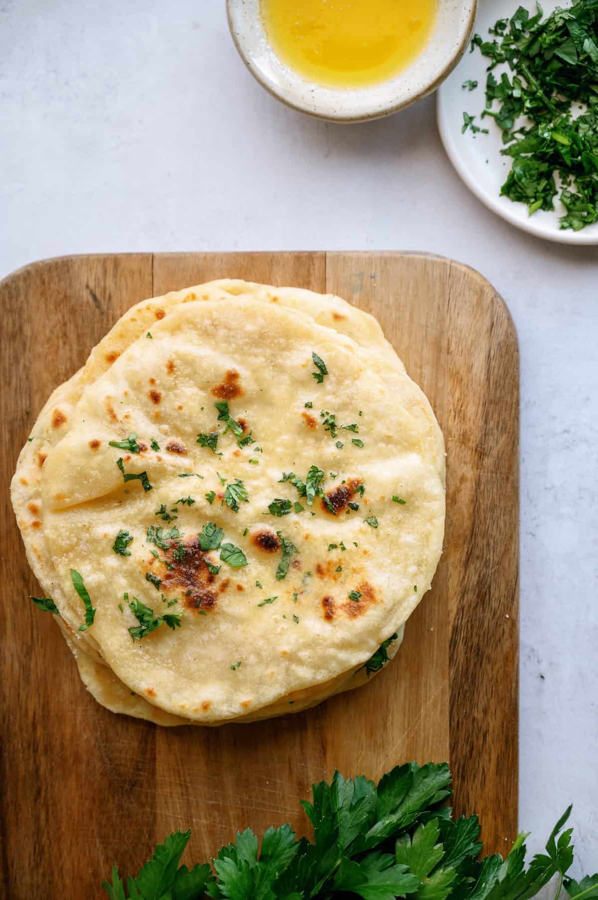 A stack of flatbreads topped with chopped herbs on a wooden board, with a bowl of melted butter and a plate of chopped herbs nearby.