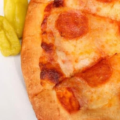 A close-up of a pepperoni pizza on a white plate with two yellow-green peppers on the side.
