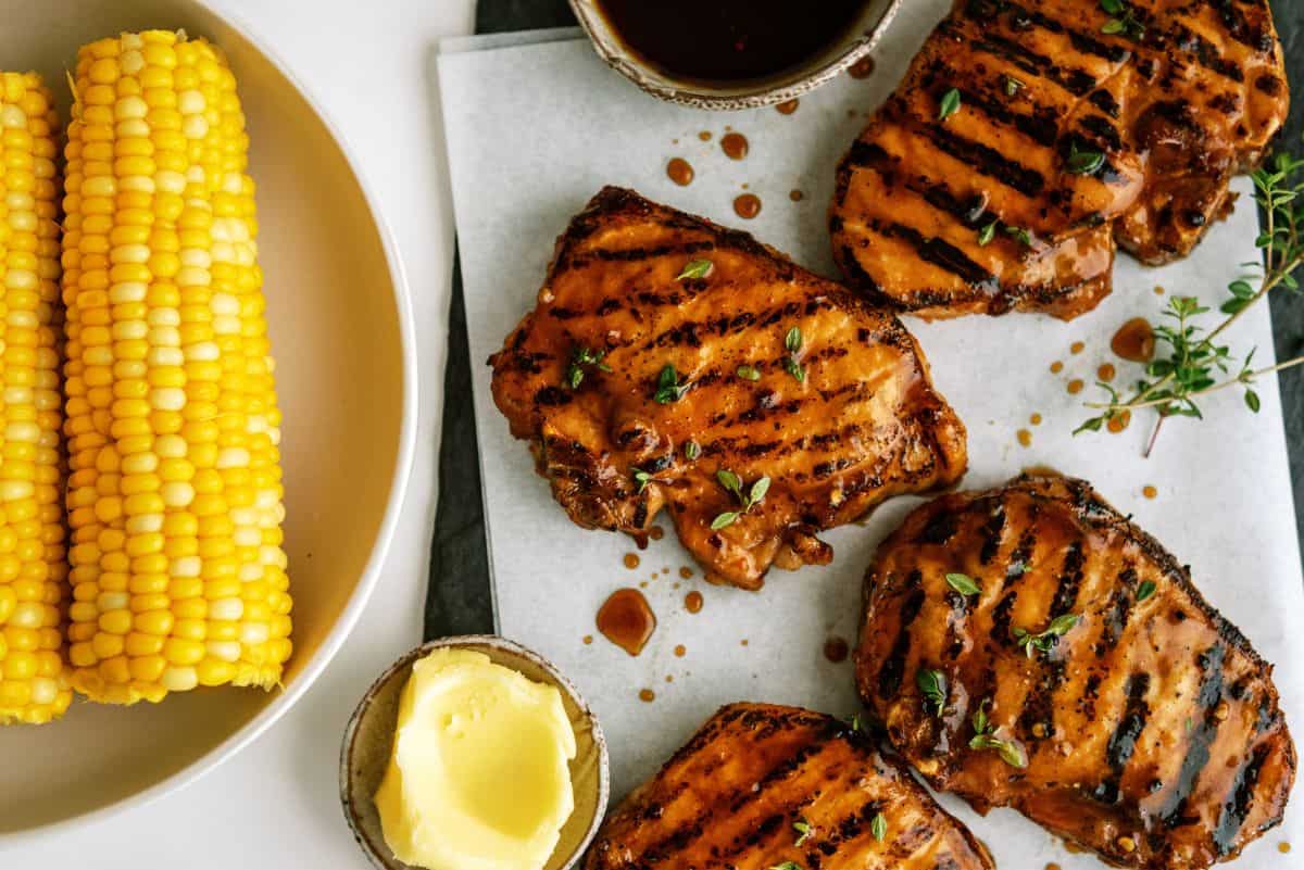Grilled pork chops garnished with herbs on a white plate, served with two pieces of corn on the cob in the background.