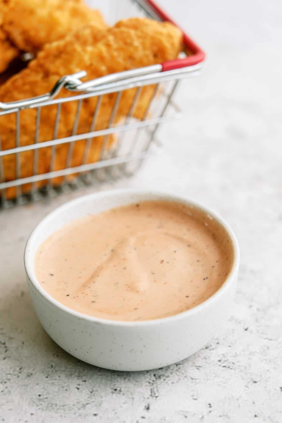 A small white bowl of creamy dipping sauce sits in front of a metal basket filled with breaded chicken tenders on a light surface.