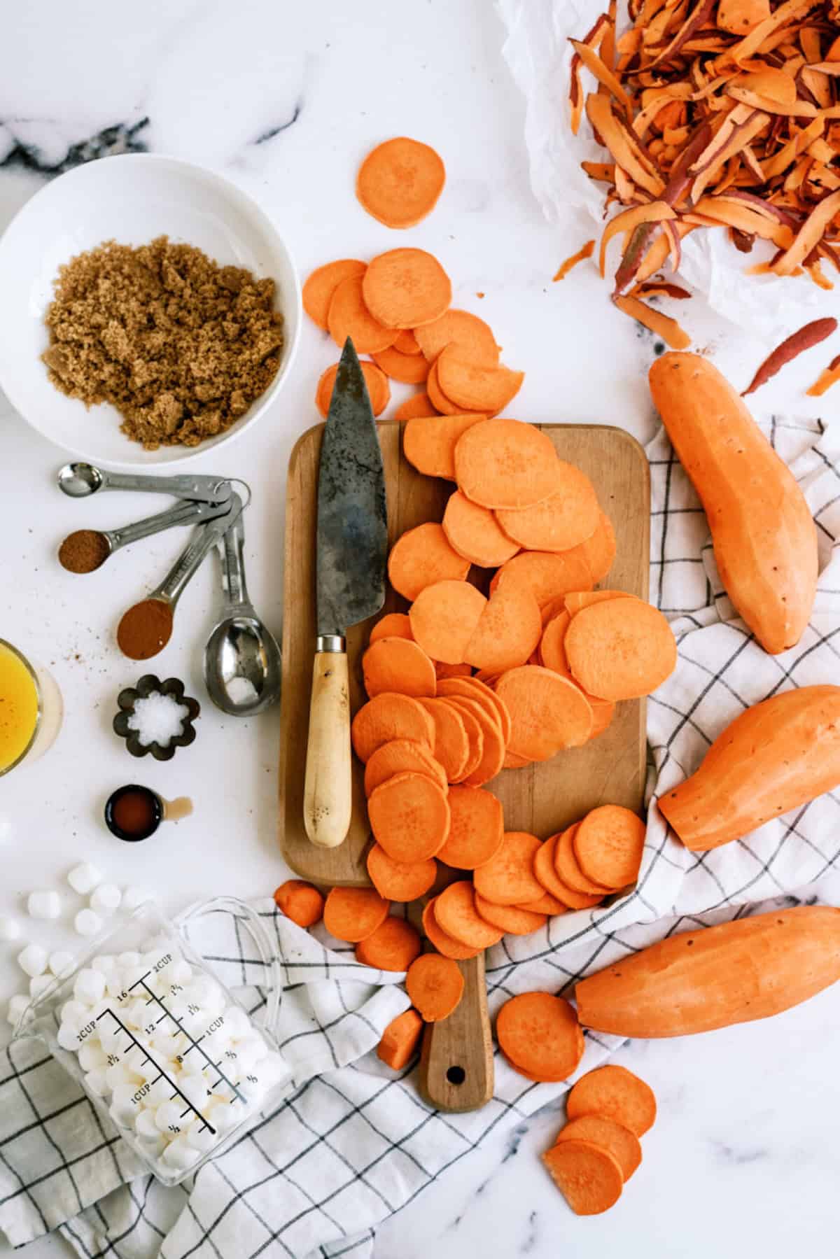 Sliced sweet potatoes on a cutting board with a knife, surrounded by measuring spoons, brown sugar, marshmallows, and sweet potato peels on a white surface.