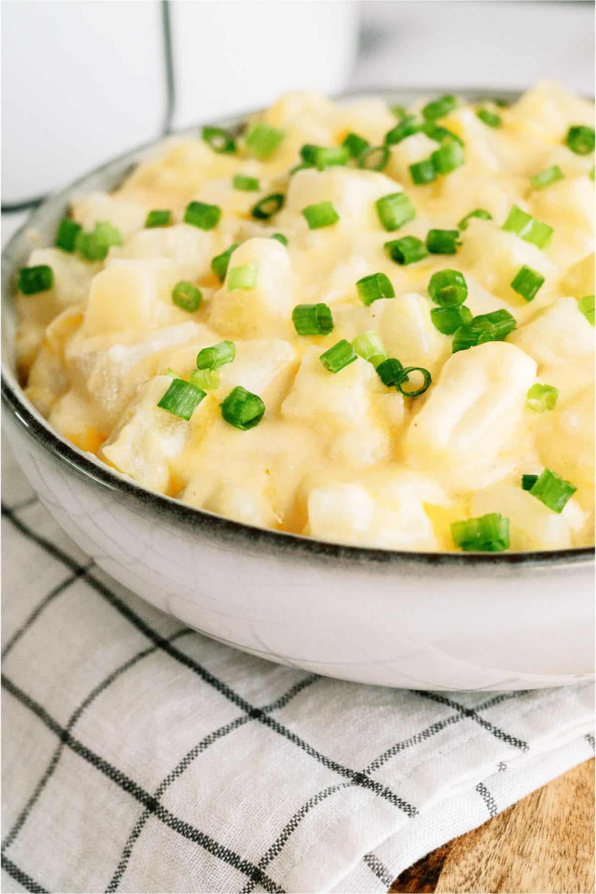 A bowl of cheesy potato casserole topped with chopped green onions, placed on a white and black checkered cloth.