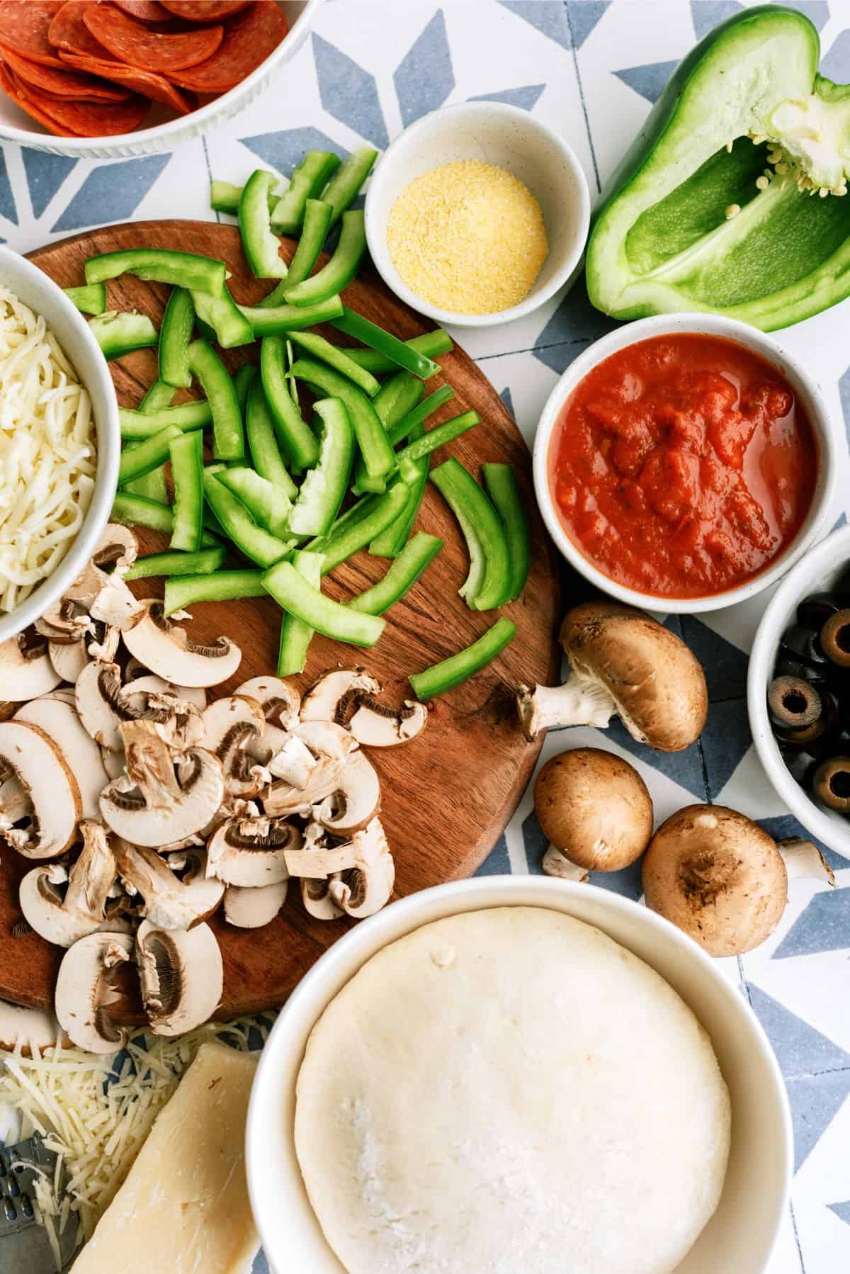 A wooden board with sliced green bell peppers and mushrooms, surrounded by pizza dough, tomato sauce, mozzarella, pepperoni, olives, cornmeal, and a halved green bell pepper.