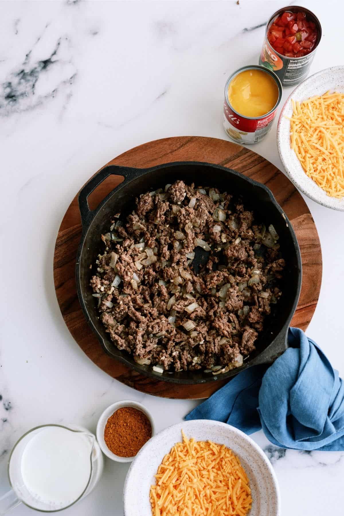 Cast iron skillet with cooked ground beef and onions on a wooden board, surrounded by shredded cheese, milk, taco seasoning, canned soup, and diced tomatoes.