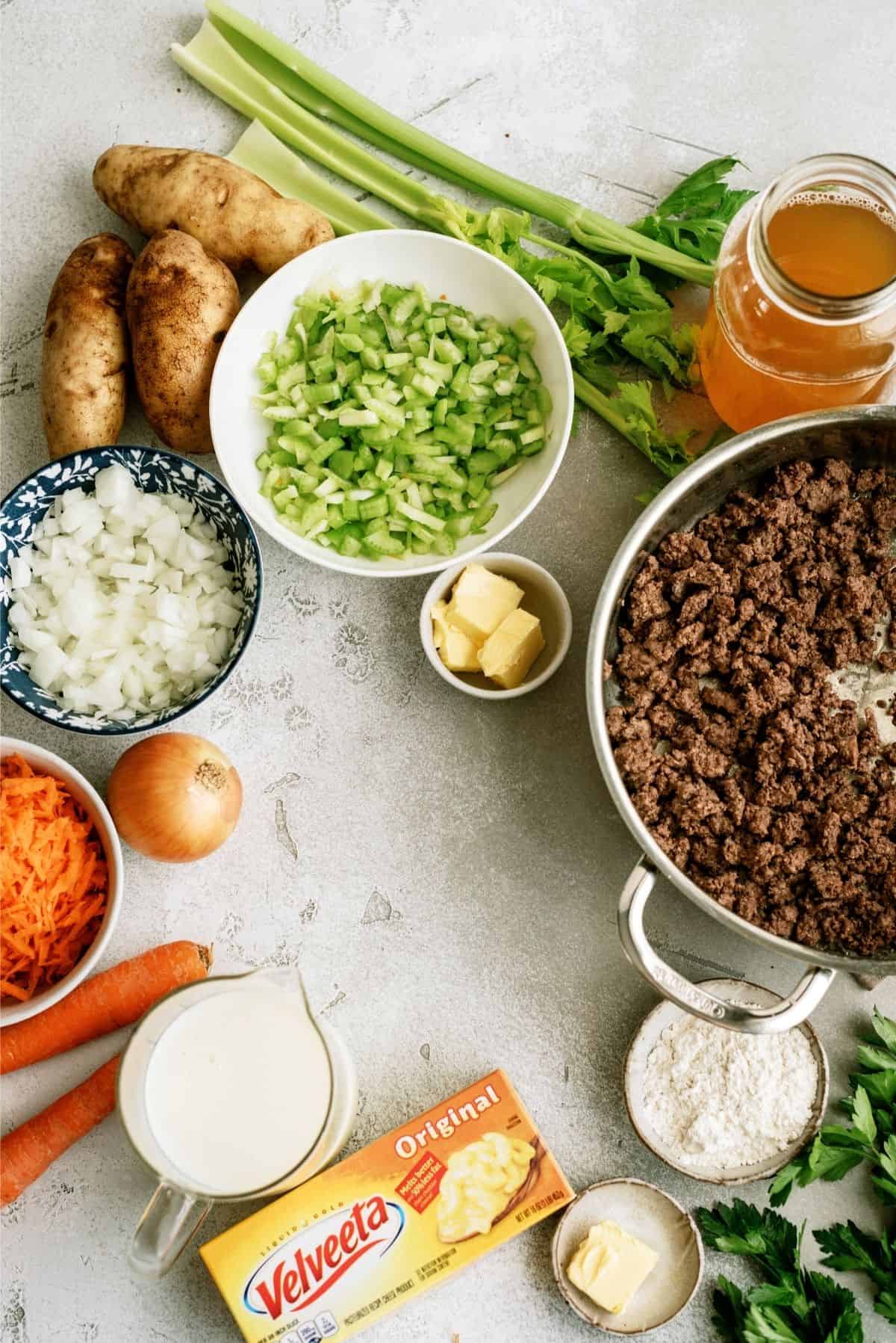 Ingredients for a soup or stew: ground beef in a pot, chopped celery, diced onion, carrots, potatoes, butter, flour, broth, Velveeta cheese, cream, and parsley arranged on a countertop.