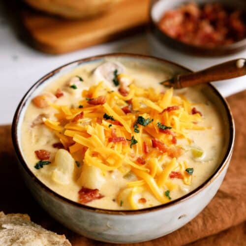 A bowl of creamy soup topped with shredded cheddar cheese, chopped bacon, and herbs, with a slice of bread on the side.