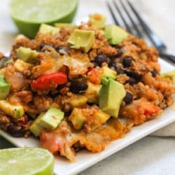 A square white plate with a serving of quinoa casserole topped with avocado chunks, surrounded by lime halves and a fork in the background.