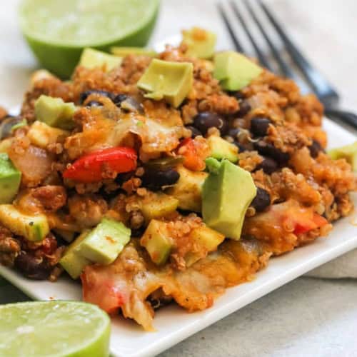 A square white plate with a serving of quinoa casserole topped with avocado chunks, surrounded by lime halves and a fork in the background.